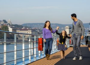 a family made of two adults and one child walking along the outside deck a ship with a building on the coast in the background