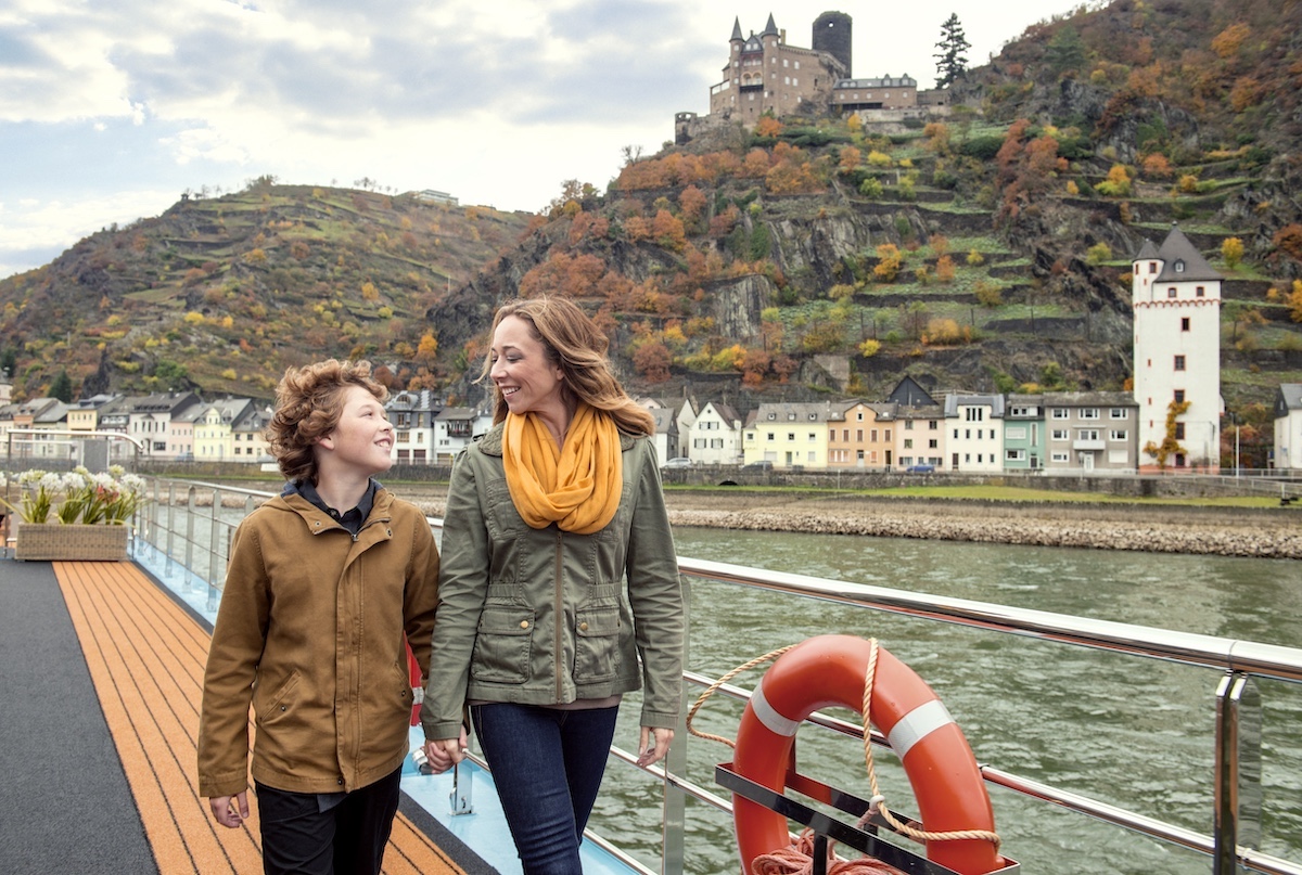a mother and son walking alongside the deck of a river cruise ship with a town in the background